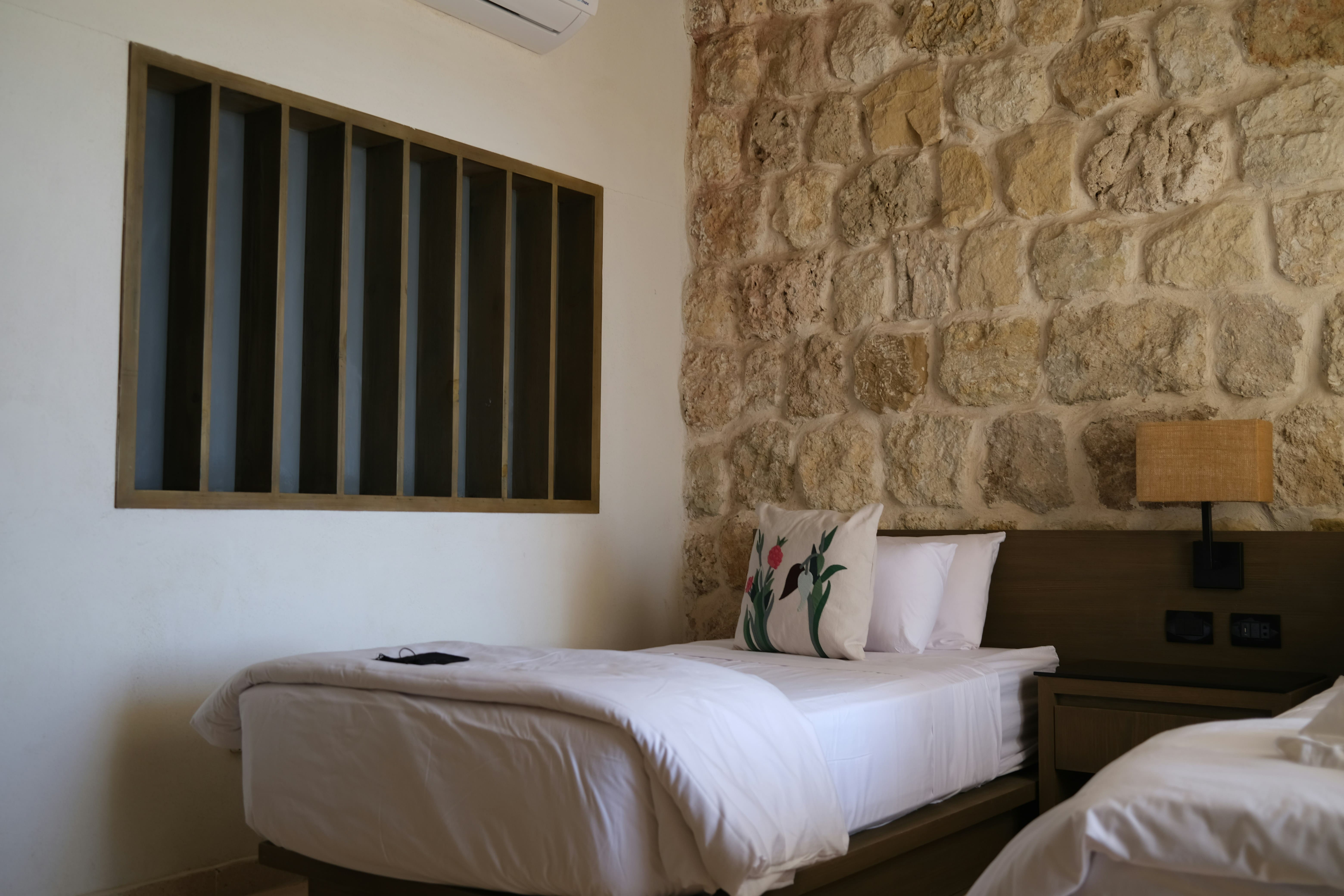 Chambre Lavande : lit aux draps blancs contre un mur en pierres apparentes, coussin brodé de fleurs et applique en raphia