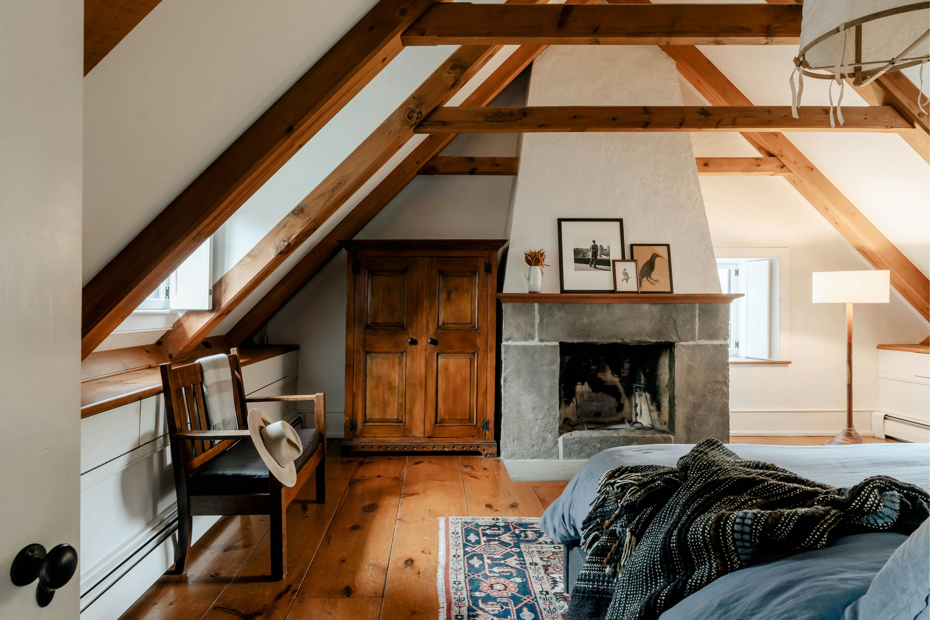 Suite Oliviers sous les toits : charpente en bois apparente, cheminée en pierre, armoire ancienne, parquet et tapis coloré au pied du lit